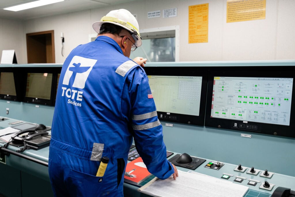 Worker in blue uniform and helmet monitoring control panels in a control room.