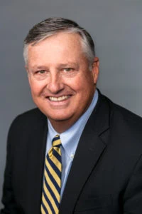 Portrait of a smiling man in a suit and striped tie against a gray background.