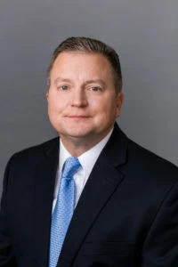 Professional portrait of a man in a suit and blue tie against a gray background.