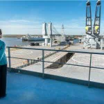 Person standing on a ship's deck with cranes and port in the background.
