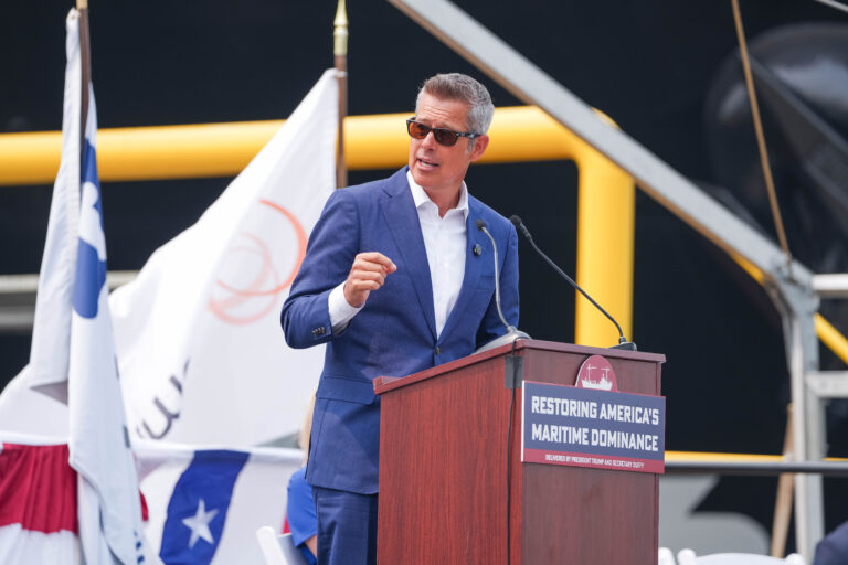 Man in blue suit speaking at podium with flags and ship in background.