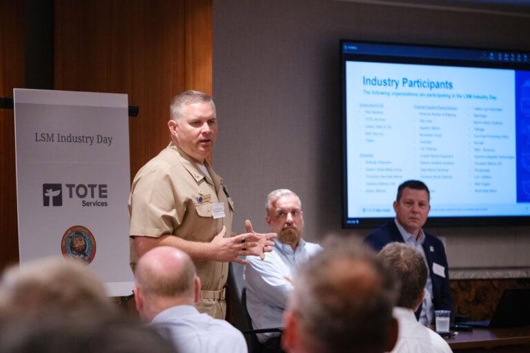 Man in tan uniform speaking at a conference with a screen and two seated men behind him.