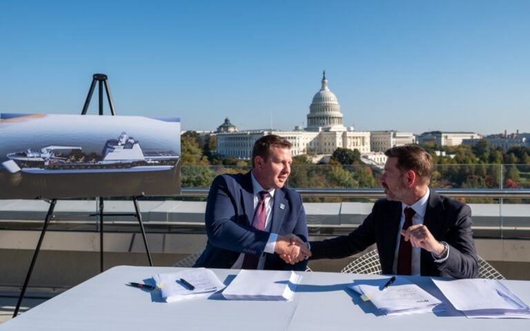 Two men in suits shaking hands at a signing table on a rooftop with a model ship and the Capitol in-.