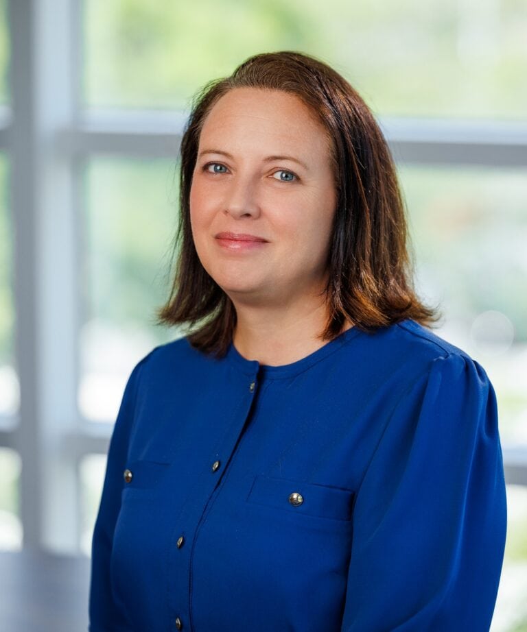 Jennifer Blakeslee in a blue blouse standing indoors with a blurred window background.
