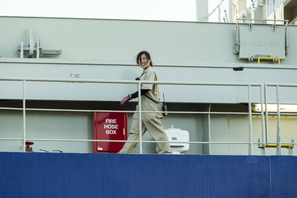 Woman in beige coveralls walking on a ship's deck near fire safety equipment.