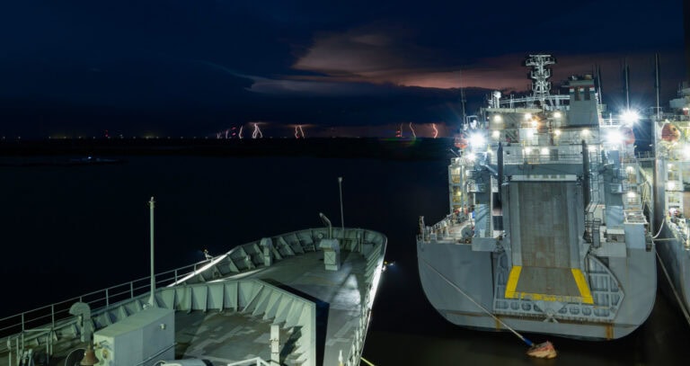 Large cargo ship docked at port during nighttime with city lights in the background.