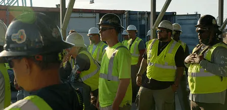 Workers in yellow vests standing on a dock with containers in background.