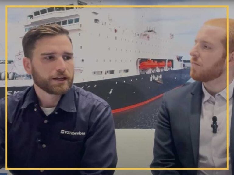 Two men discussing vessel construction in front of a large ship model.