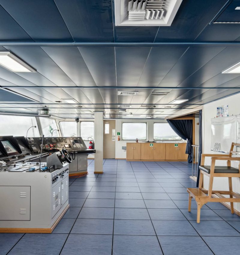 The image shows the interior of a ship's control room or bridge with navigation and communication eq.