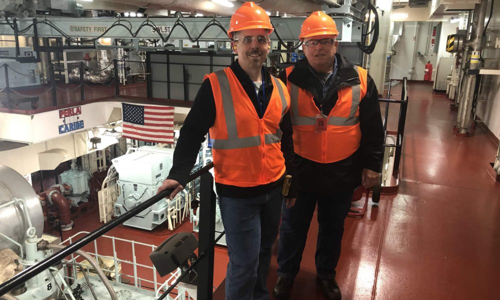 Two men in orange safety vests and helmets standing on an industrial platform.