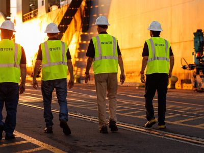 Ketterman_250729_3129-web Five workers in safety vests and helmets walking along a shipyard at sunset.