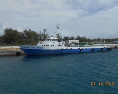 USNS FAST TEMPO ship docked at a pier with trees and buildings in the background, under a cloudy sky.