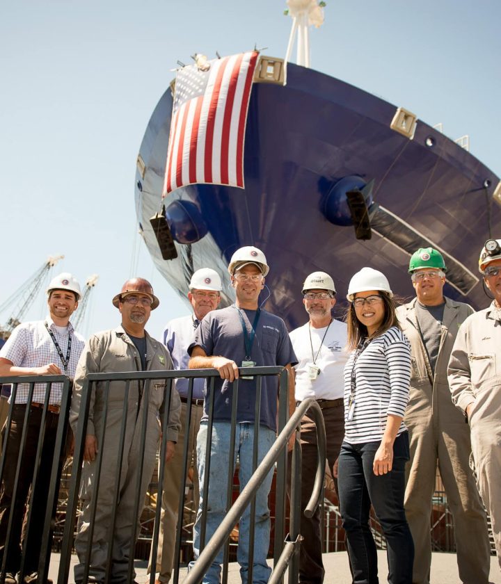 US flag on ship's bow with group of engineers and workers at dockyard.
