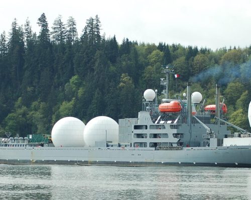 Aerial view of a large military ship with radar domes sailing in a forested area.