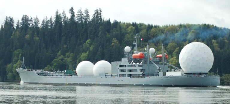 Aerial view of a large military ship with radar domes sailing in a forested area.
