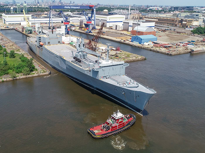 Large gray fast sealift ship docked at a port with cranes and industrial buildings in the background.